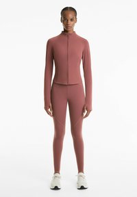 Žena stojaca oblečená v zladenom mauve zip-up saku a legínach, s bielymi športovými topánkami na bielom pozadí.