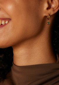 Goldene Creole mit einem kleinen Türkisstein, der von der Creole hängt, präsentiert auf texturierter Haut nahe der Kieferlinie.