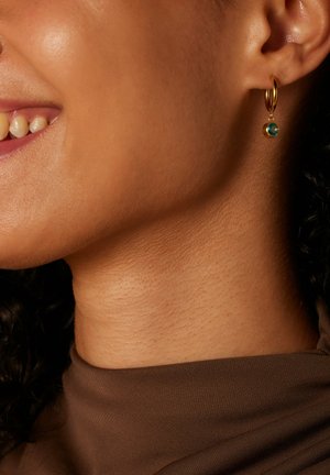 Boucle d'oreille en or présentant une petite pierre turquoise, suspendue à la boucle, mise en valeur sur une peau texturée près de la mâchoire.