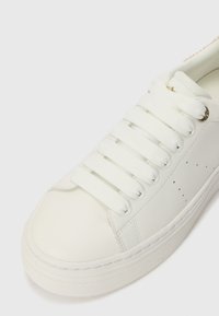 Sneaker bianco realizzato in materiale sintetico liscio, con lacci bianchi piatti e sottili perforazioni. Punta arrotondata con cuciture a contrasto e suola in gomma.