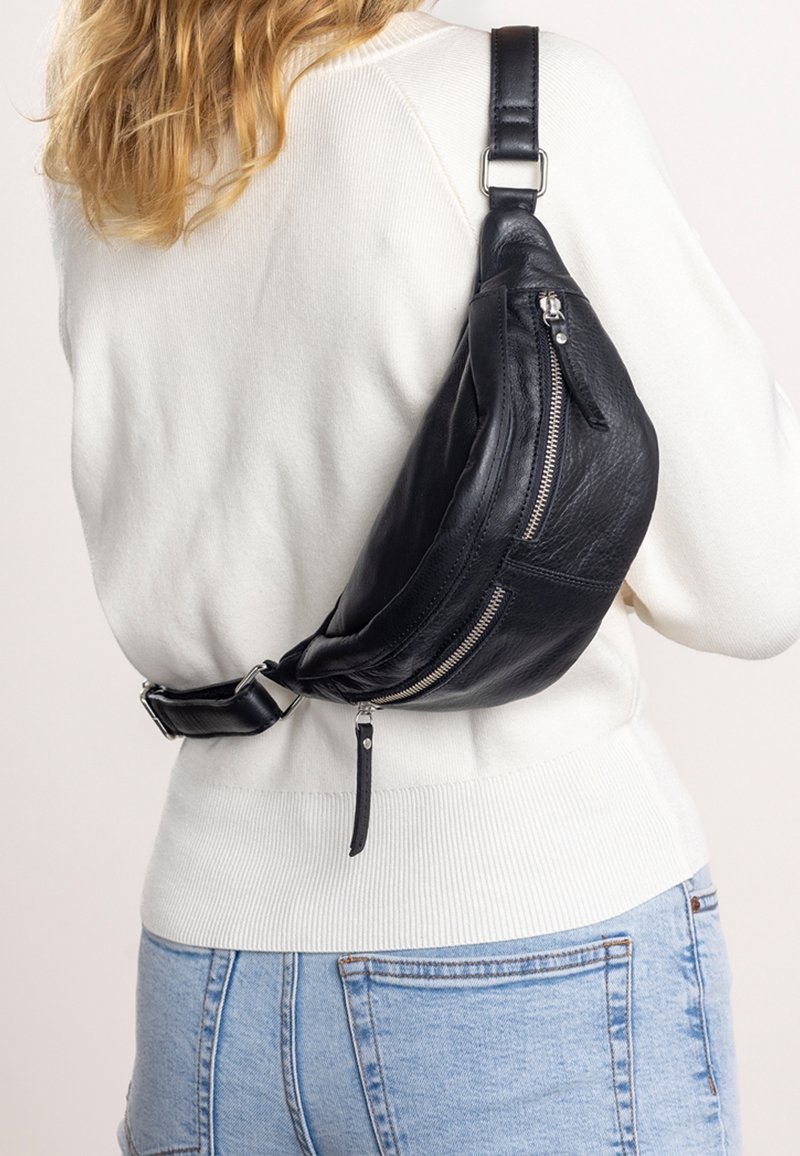 Femme portant un sac bandoulière en cuir noir avec des fermetures éclair argentées sur un pull blanc et un jean en denim bleu clair.