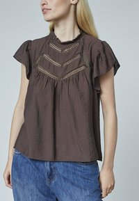 Brun bluse med korte, flæsekantede ærmer, blonde detaljer i et chevronmønster og en afslappet pasform. Blødt, tekstureret stof.