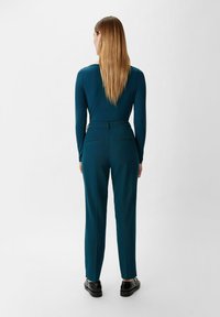 Tealowa bluzka z długim rękawem zestawiona z dopasowanymi spodniami prostymi. Spodnie mają boczne kieszenie oraz gładką teksturę. Czarnym butom towarzyszy całość stylizacji.