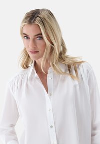 Camicia bianca con bottoni, caratterizzata da colletto, maniche lunghe con dettagli arricciati e una leggero lucido; realizzata in tessuto leggero e liscio.