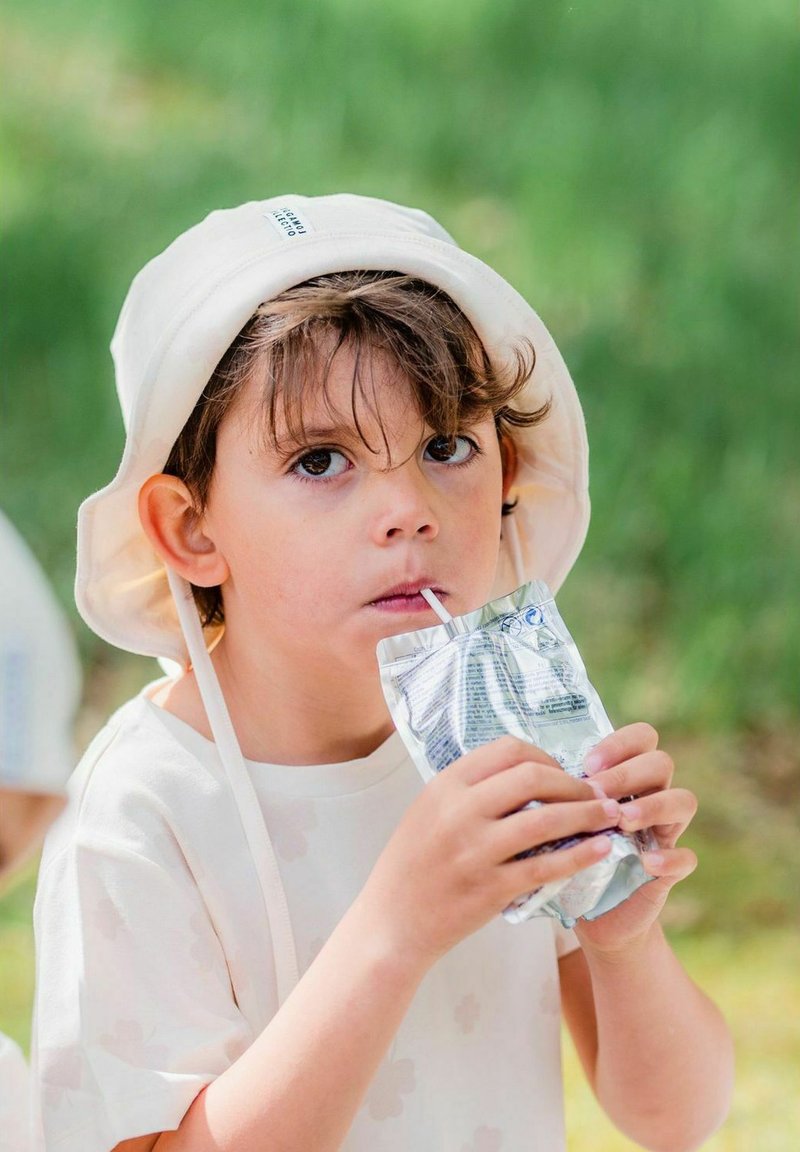 Lapsi, joka on pukeutunut vaaleaan ämpärilakkiin ja valkoiseen puseroon, pitää hopeista mehujuomapussukkaa, jossa on pilliruisku, sumean vihreän taustan edessä.