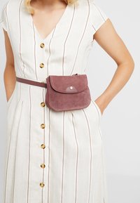 Zamszowa torebka noszona na krzyż w odcieniu mauve z zaokrąglonym klapą, ozdobiona złotą zapięciem na ćwiek i regulowanym paskiem, noszona na tle paskowanej sukienki.