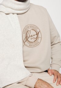 Beige mikina má embosované logo v hnědé barvě, je doplněná světle béžovým pleteným šálkem, což ukazuje na jemné textury a jednoduchý, moderní design.