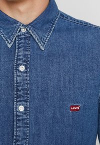 Denim overhemd in middenblauw met een button-down kraag. Voorzien van witte knopen en een rood Levi's logo op de linkerbovenborst.