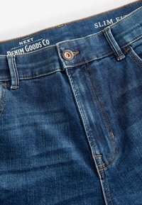 Slim-fit denim jeans in donkerblauw, met een knoopsluiting en contrasterend oranje stiksel. De stof heeft een fijne textuur en een subtiele vervaging.