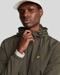 Junger Mann in einer olivgrünen Jacke mit Reißverschluss und einem gelben Vogel-Logo sowie einer weißen Kappe, der den Kragen der Jacke anpasst.