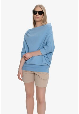 Kobieta w czarnych okularach przeciwsłonecznych, luźnej jasnoniebieskiej bluzce z rękawami do łokcia i beżowych szortach, stojąca na białym tle.