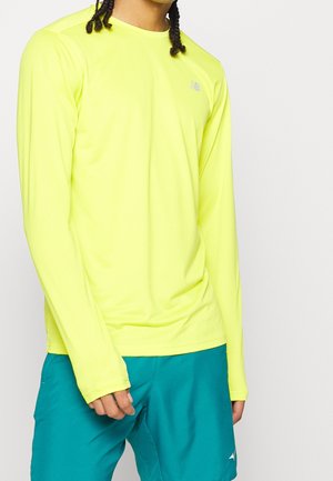 Persona che indossa una maglia sportiva a maniche lunghe di colore giallo brillante e pantaloncini sportivi turchese su sfondo bianco uniforme.