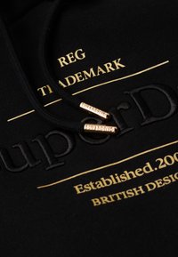 Sudadera negra con texto y bordado en dorado. Cordones con aglets metálicos grabados "SUPERDRY". Textura suave, diseño informal.