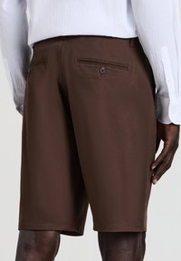 Un short marron en tissu lisse présente deux poches arrière avec des boutons et un design droit. Une chemise blanche à boutons est visible au-dessus.