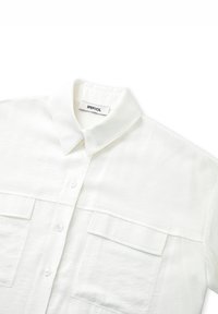 Chemise blanche à col boutonné, fabriquée en tissu léger, avec deux poches sur la poitrine et une finition texturée. L'étiquette indique "IPEKYOL."