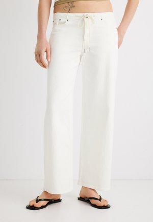 Személy, aki magas derekú, fehér, széles szárú nadrágot visel húzózsinórral és fekete flip-flop papucsot, egy egyszerű fehér háttér előtt áll.