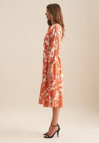 Orangenes, blumenmotiviertes knielanges Kleid mit langen Puffärmeln, tailliertem Mittelteil und schwingendem Rock. Getragen mit schwarzen High-Heels.