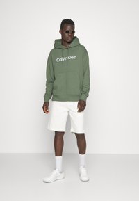 Felpa verde oliva con logo bianco "Calvin Klein", tasca frontale e cappuccio con coulisse; abbinata a pantaloni corti bianchi e sneaker bianche.