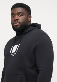 Sudadera negra de algodón suave, con un diseño de logo en blanco en la parte frontal y un bolsillo tipo canguro. Incluye capucha con cordones y un ajuste relajado.