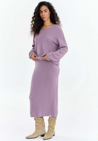 Fioletowa dzianinowa sukienka o luźnym kroju, z długimi rękawami i zaokrąglonym dekoltem. Miękka w dotyku, noszona z beżowymi kozakami sięgającymi kolan.