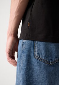 T-shirt en coton noir avec une petite étiquette orange 'BOSS', associé à un jean en denim bleu clair, avec des détails de couture sur les poches.
