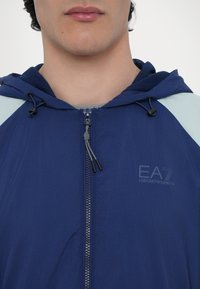 Navy blue zip-up jacket with a hood, featuring light blue shoulder accents, a textured fabric, and branded logo on the chest.