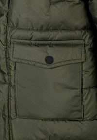 Olivnozeleni pufer jakna z teksturo, ki ima kvadratni vzorec, z zaščitno žepom zaklenjenim s črnim gumbom na levi strani.