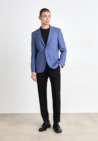 Blazer bleu deschis cu un singur nasture, rever cutat și buzunare frontale, asortat cu pantaloni negri și pantofi negri, pe un fundal neutru.