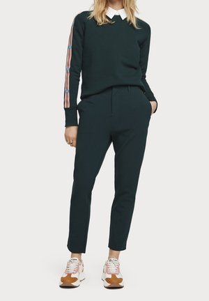 Kobieta w ciemnozielonym topie z długimi rękawami w paski, dopasowanych spodniach, białej koszuli z kołnierzykiem pod spodem oraz białych brązowych trampkach.