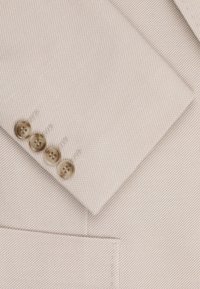 Beżowa tkanina marynarki z teksturowanym splotem. Cztery jasnobrązowe guziki z czterema otworami, przyszyte do mankietu z ukośnym klapem.