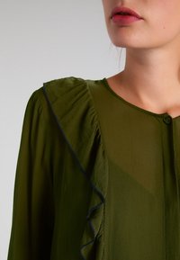 Blusa verde oliva con spalla sinistra arricciata e bordi blu. Presenta chiusura con bottoni e un tessuto leggero e trasparente.