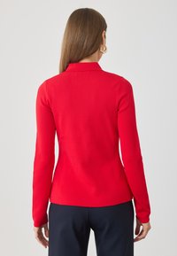 Mujer con el pelo castaño liso, lleva pendientes de aro dorados, una blusa roja de manga larga y pantalones oscuros, de pie mirando hacia afuera contra un fondo sencillo.