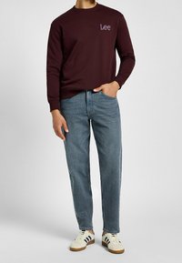 Uomo che indossa una felpa maroon Lee, jeans blu con una mano in tasca e sneakers bianche con strisce nere su sfondo grigio.