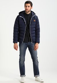 Navy puffer jacket with a fitted design, white zippers, and a hood. Worn over a black shirt and paired with blue jeans and white sneakers.