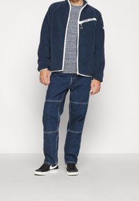 Fleece bunda v námořnické modři s bílým zipem a akcenty, kombinovaná s modrými denimovými kalhotami s vyšívanými detaily a černými teniskami.