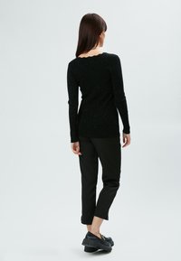 Schwarzer Pullover mit langen Ärmeln, gewelltem Ausschnitt und strukturierter Oberfläche. Kombiniert mit schwarzen Hosen und klobigen, schwarzen Schuhen.