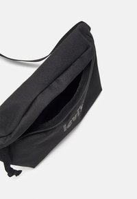 Borsa a tracolla nera in tessuto con zip, caratterizzata da una superficie testurizzata e un logo grigio chiaro ben visibile. Design compatto con tracolla regolabile.