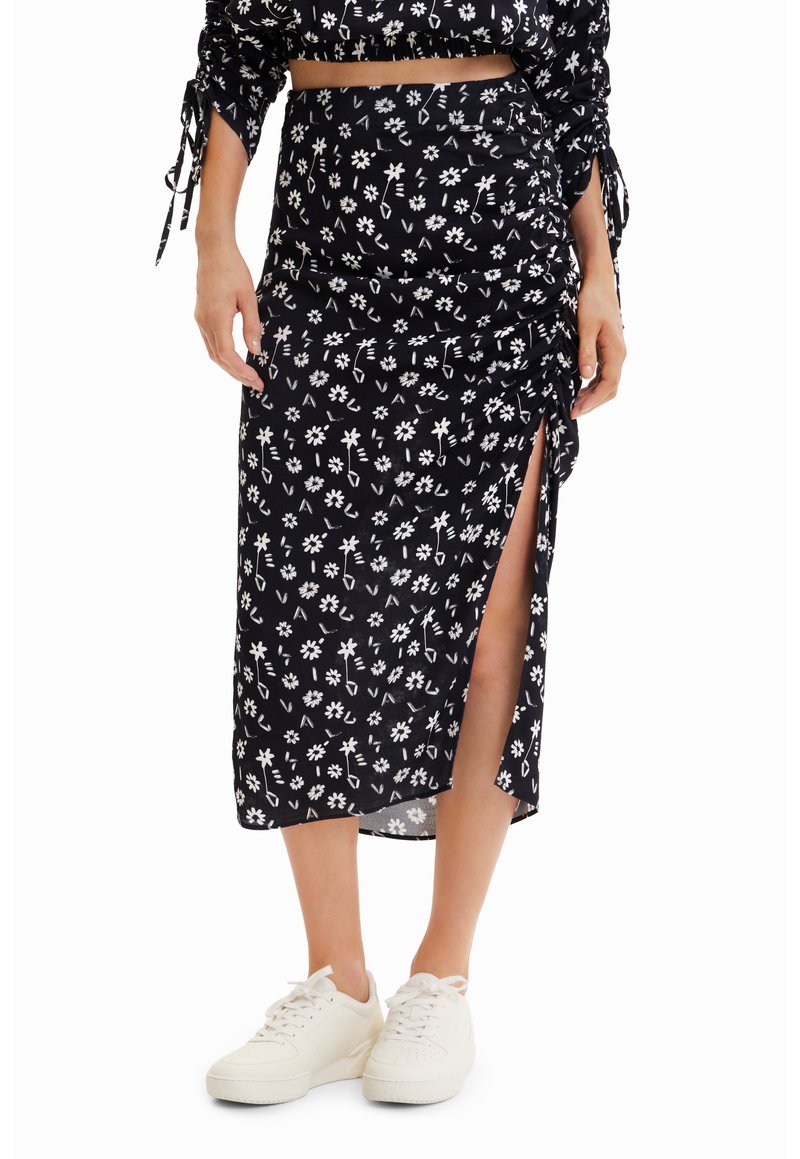 Jupe noire mi-longue avec motif floral blanc, fente latérale et détail froncé portée avec des baskets blanches.