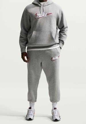 Mężczyzna w szarej bluzie z kapturem i spodniach dresowych Nike z białym i czerwonym logo, białych skarpetkach i białych butach sportowych, stojący na jednolitym tle.