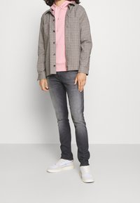 Šedá košile s kostkovaným vzorem, pod ní růžová mikina, úzké tmavé džíny a bílé tenisky. Oblečení zahrnuje vrstvené textury a barvy.