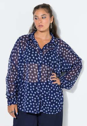 Frau trägt eine transparente marineblaue Bluse mit weißen Punkten, vorne geknöpft und langen Ärmeln, posiert mit einer Hand auf der Hüfte.