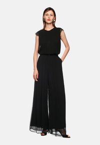 Schwarzer Jumpsuit mit fließender Silhouette, transparenter Ärmel, gefältelten Details, weitem Bein und Seitentaschen. Kombiniert mit schwarzen High-Heels.