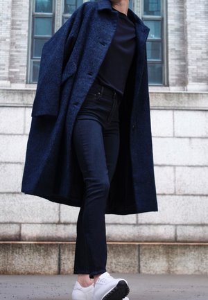 Personne portant un manteau bleu foncé, une chemise noire, un jean foncé et des baskets blanches, debout contre un bâtiment en pierre avec des fenêtres.