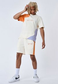 Sudadera de manga corta color crema con detalles en naranja y lavanda, combinada con pantalones cortos color crema que tienen una franja naranja y un logotipo.