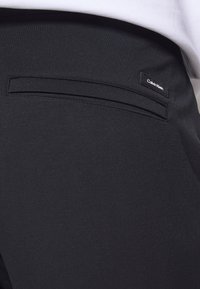 Gros plan de la poche arrière d'un pantalon noir avec une étiquette Calvin Klein et une chemise blanche rentrée au-dessus de la ceinture.