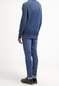 Mörkblå stickad tröja med ett fläckigt mönster, kombinerad med ljusblå denimjeans. Modellen bär svarta skor. Bakifrån.