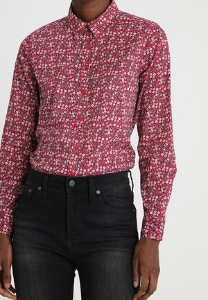 Rotes langärmeliges Hemd mit Knöpfen und kleinem weißem Blumenmuster, in hoch taillierte schwarze Jeans mit Vordertaschen gesteckt.