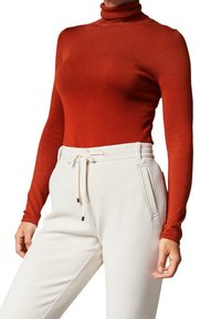 Pull à col roulé rouge à manches longues associé à un pantalon blanc texturé avec taille à cordon et poches latérales.