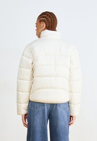 Kremfarget pufferjakke med horisontale sømmer, høy krage og elastiske mansjetter, sammen med vide denimjeans.