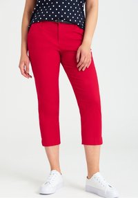 Pantalon capri rouge à coupe ajustée, avec des revers roulés. Associé à un haut bleu marine à pois et des baskets blanches.